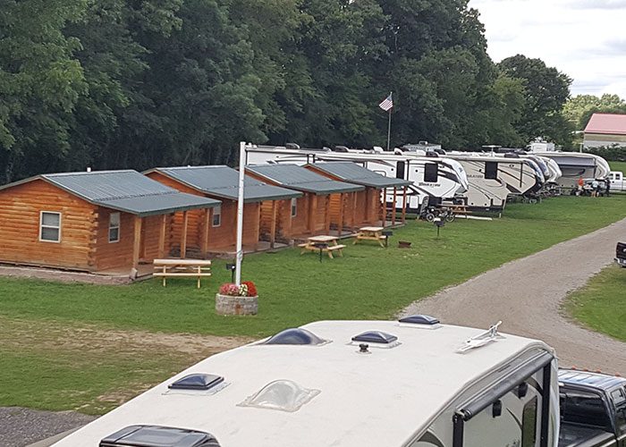 Row of small log cabins with picnic tables and a flagpole, beside parked RVs
